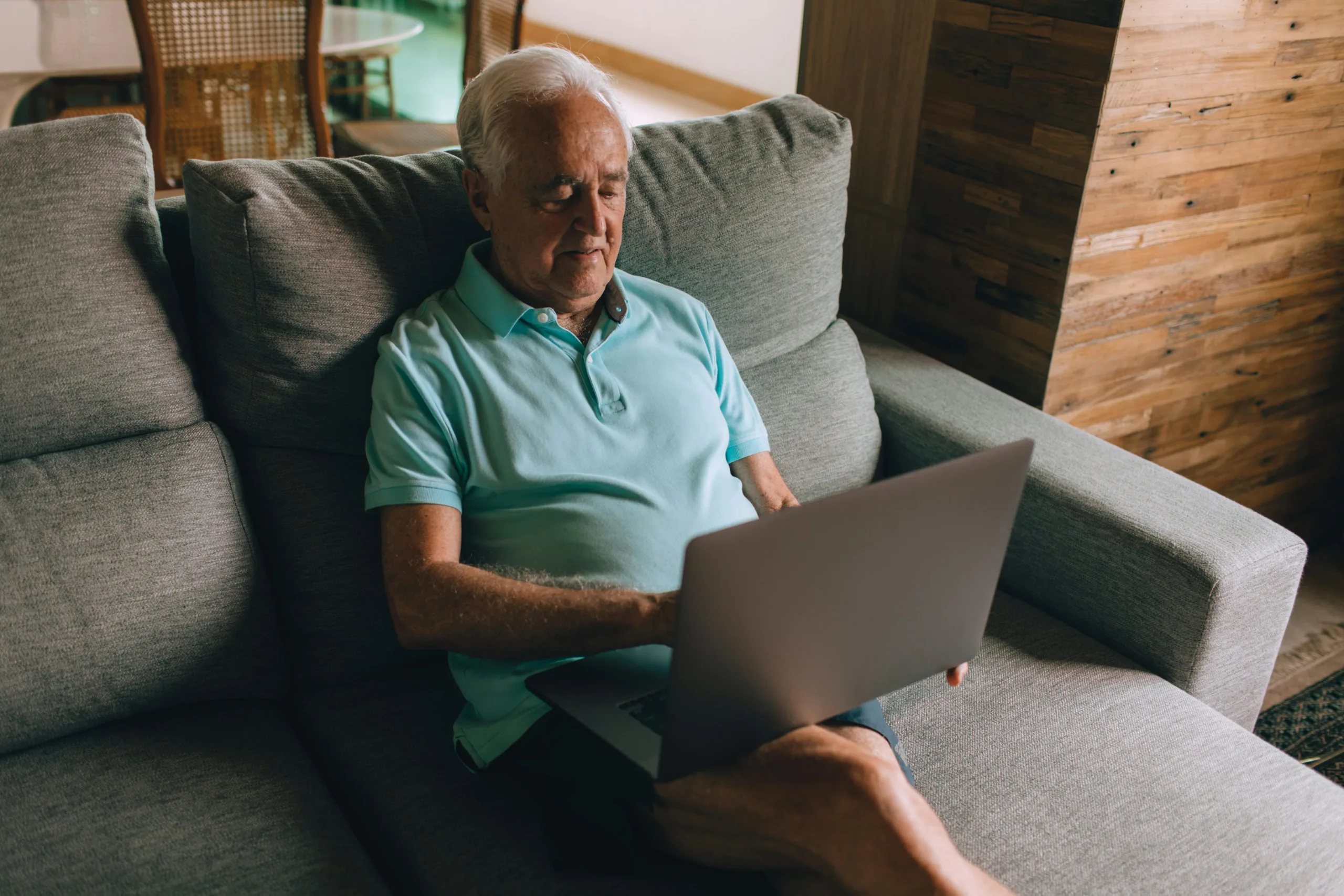 Senior man in blue polo shirt using a laptop while sitting on a cozy couch indoors.